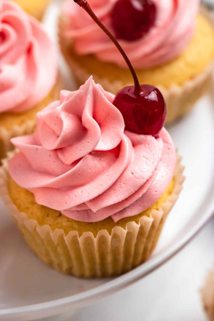 yellow colored cupcakes with pink frosting and a cherry sitting on the side of cupcake 