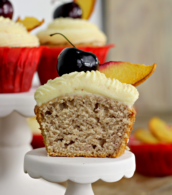 a peach cupcake cut in half with cherry and peach slice on top of frosting. The cupcake is sitting on a cupcake stand 