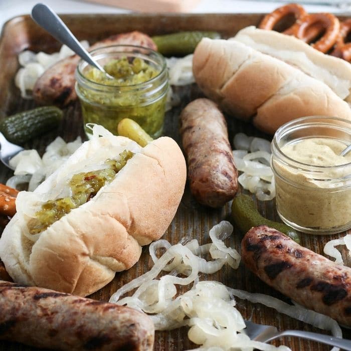 grilled brats on cooking sheet with condiments all around it 