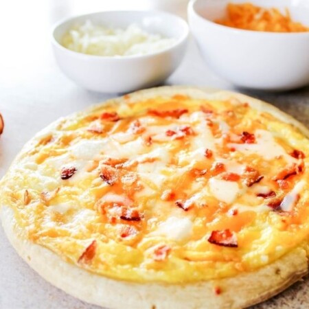 breakfast pizza on wooden cutting board with cheese in bowls and bacon beside it
