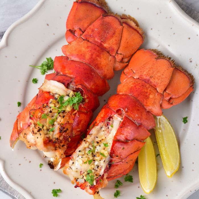 two lobster tails on plate drizzled with herbs and lemon wedges on plate 