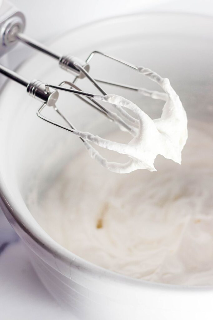 whipped cream sitting on the mixer attachments over a white bowl with whipped cream in it