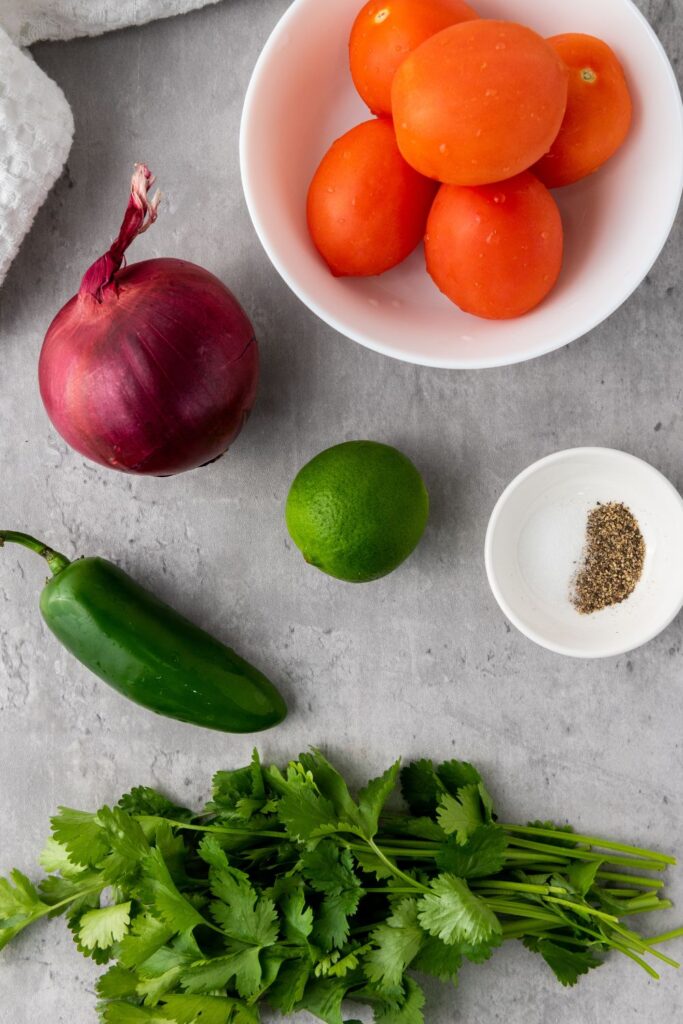 ingredients for salsa on table. Tomatoes, red onion, jalapeno, lime, cilantro, and salt and pepper