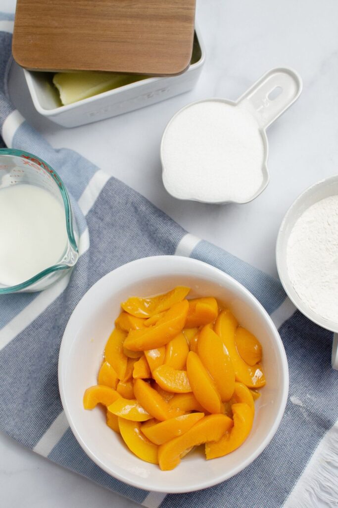 peaches in a bowl, flour, sugar and milk in bowls around the peaches on table