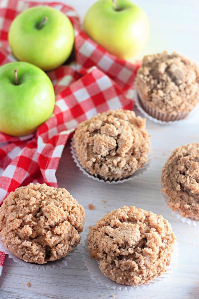 five muffins on table with cloth and apples behind it