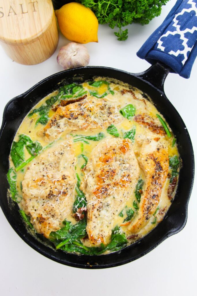 chicken Tuscan in pan on table with fresh herbs, lemon, garlic and salt sitting by the pan