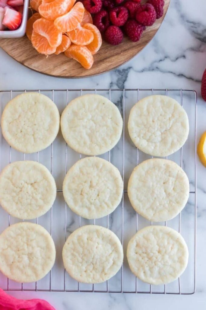 sugar cookies on a cooling rack with sliced fruit behind it on a table