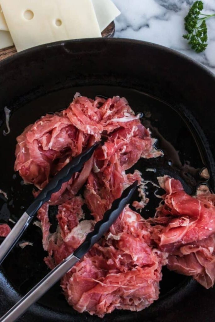 shaved steak in a cast iron skillet with tongs in pan