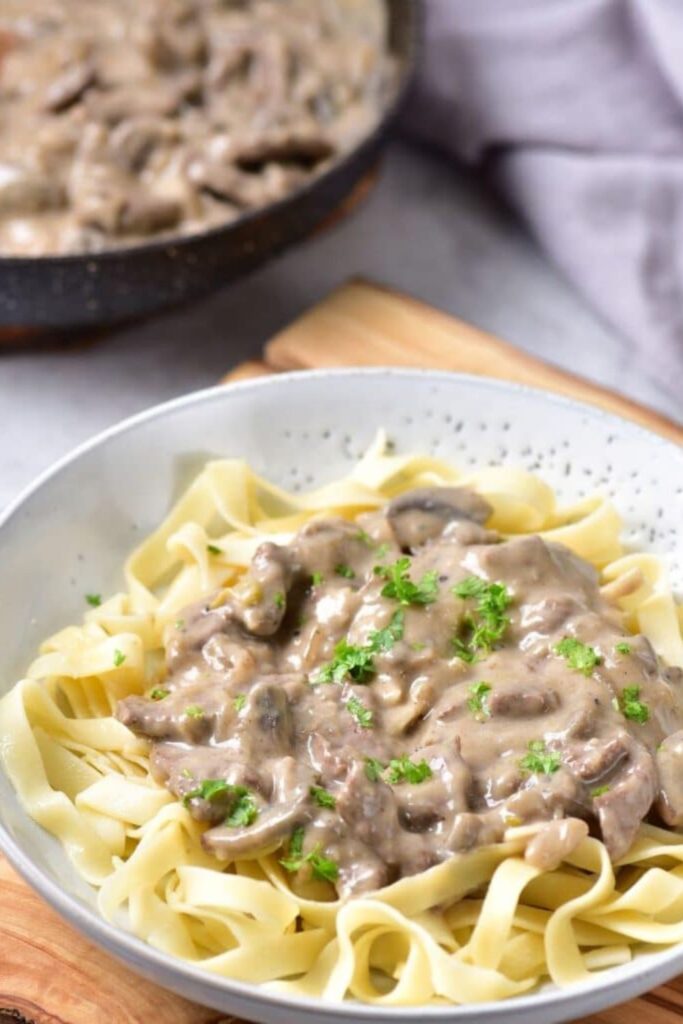 Beef Stroganoff on top of a bowl of noodles with a pan full of stroganoff in a skillet to the side