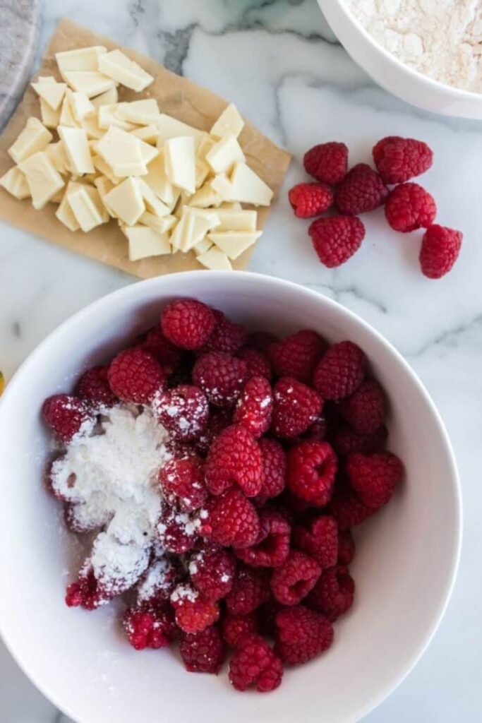 white bowl full of fresh raspberries with more on table and white chocolate on cutting board