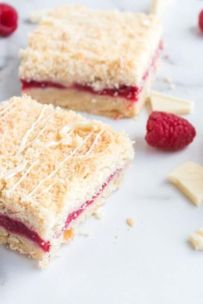 Sliced Raspberry Crumble Bars spread out on marble counter with fresh raspberries around the bars and chopped white chocolate