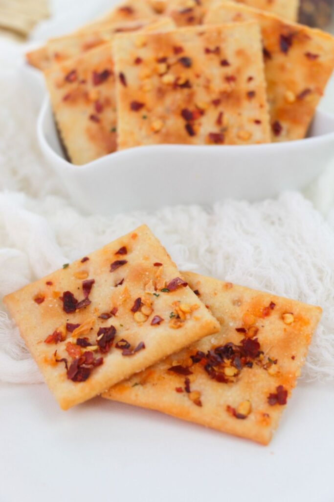 Alabama Heat Crackers in a white bowl on table with two crackers in front that are seasoned with red pepper flakes and other spices
