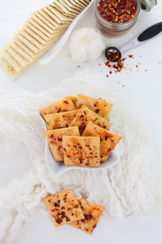 Alabama fire saltine crackers in a bowl on table with a saltine packet behind it and peppers in a measuring spoon to side