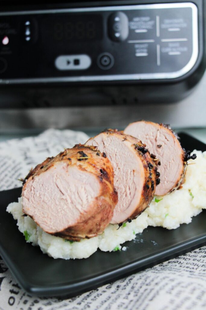 Air Fryer Pork Tenderloin sliced and placed on a plate with mashed potatoes and an air fryer behind it on the table