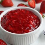 Homemade strawberry sauce in a white bowl with strawberries scattered around the table