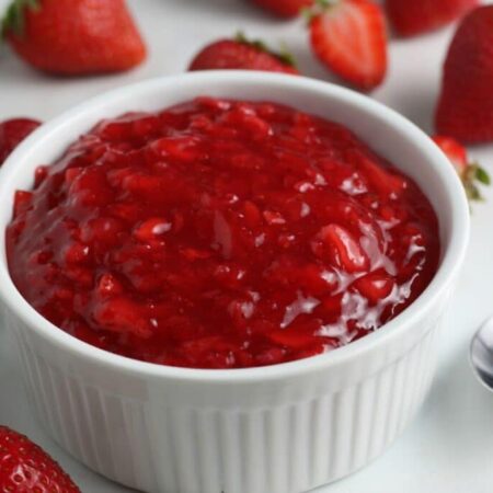 Homemade strawberry sauce in a white bowl with strawberries scattered around the table
