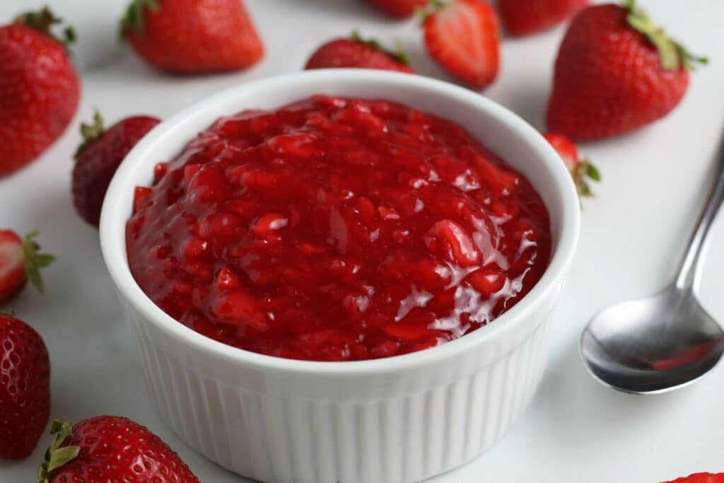 Homemade strawberry sauce in a white bowl with strawberries scattered around the table