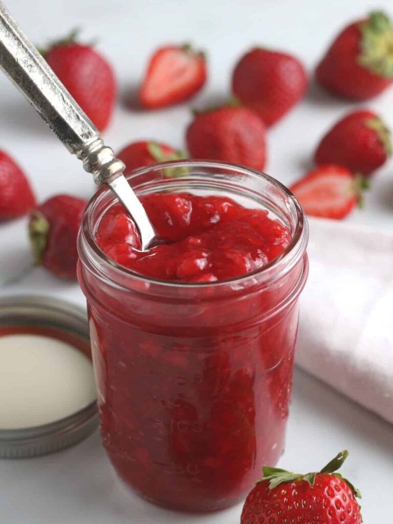 Homemade strawberry sauce in a glass jar with strawberries behind it on the table
